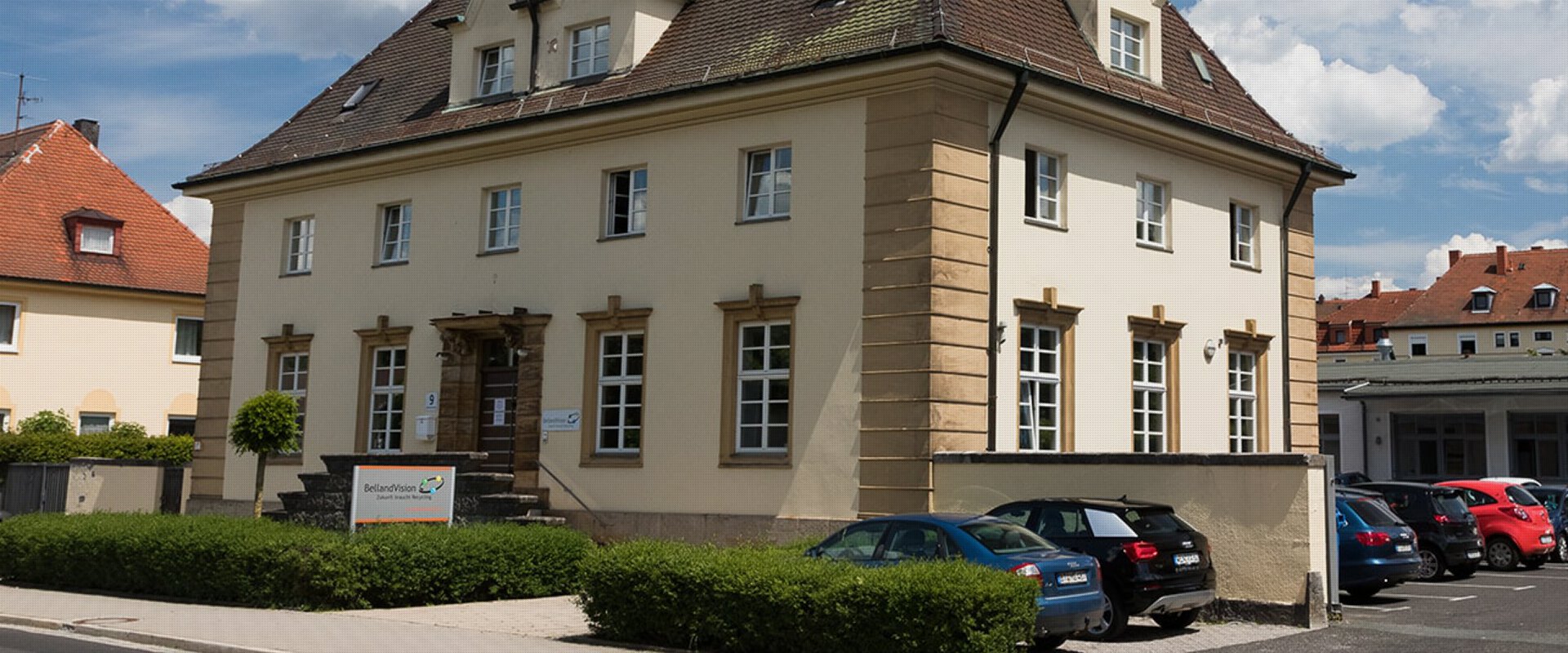 Ein historisches, zweistöckiges, sandfarbenes Gebäude mit einem dunklen Dach und heller Umrandung der Fenster zeigt den Unternehmensstandort von BellandVision in Pegnitz. 