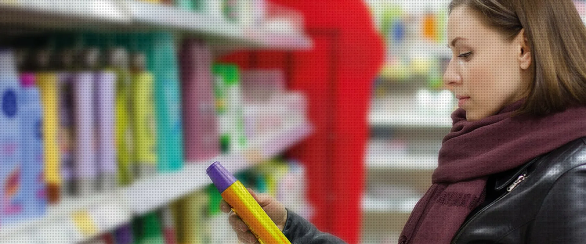Eine Frau mit einem Schal und einer Lederjacke betrachtet aufmerksam ein Produkt in ihrer Hand, während sie in einem Geschäft mit Regalen voller Körperpflegeprodukte steht.