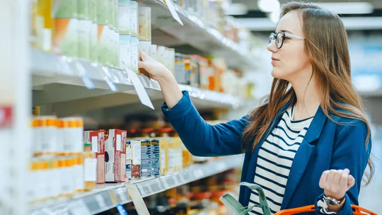 Eine junge Frau steht in einem Supermarkt und wählt sorgfältig Produkte aus einem Regal aus, während sie einen Einkaufskorb am Arm hat. Sie konzentriert sich auf die Etiketten und Preise der Produkte.