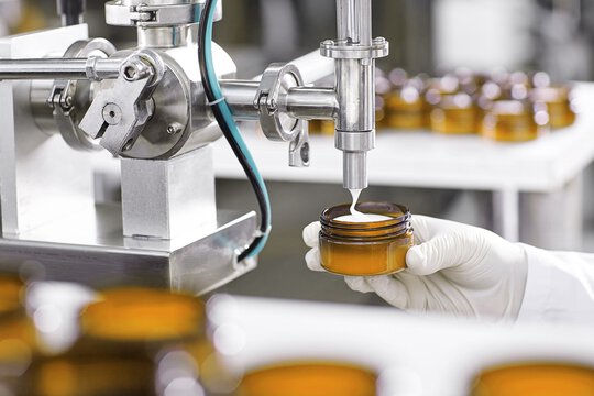 Eine Hand mit weißem Handschuh und in Laborbekleidung füllt ein Glasgefäß mit einer Maschine in einer Produktionslinie.