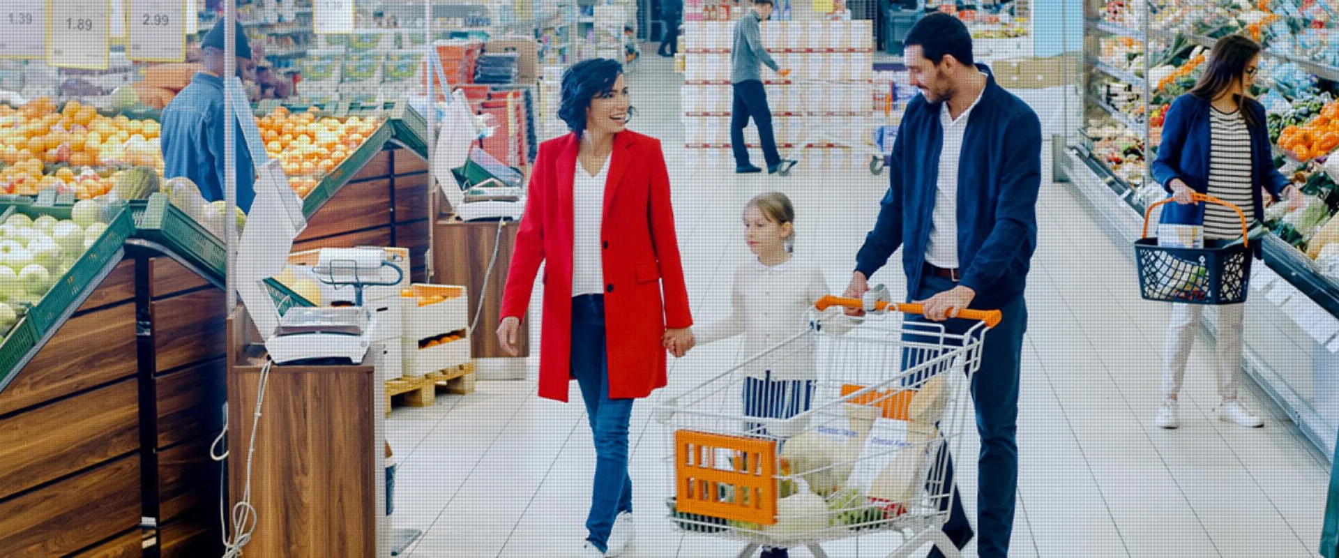 Eine Familie mit einem Kind und einem Einkaufswagen in einem hellen Supermarkt, umgeben von frischem Obst und Gemüse.