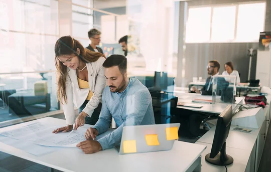 Zwei Kollegen, eine Frau und ein Mann, besprechen  Dokumente auf einem Schreibtisch in einem modernen Büro, während andere Kollegen im Hintergrund arbeiten.