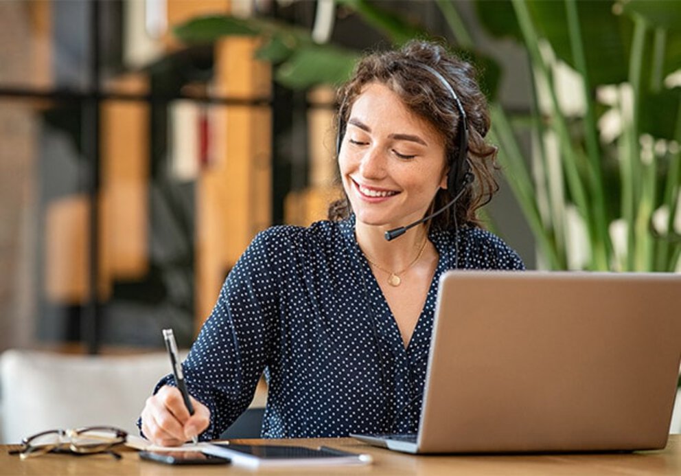 Eine freundliche Frau mit Headset sitzt vor ihrem Laptop und macht sich Notizen.