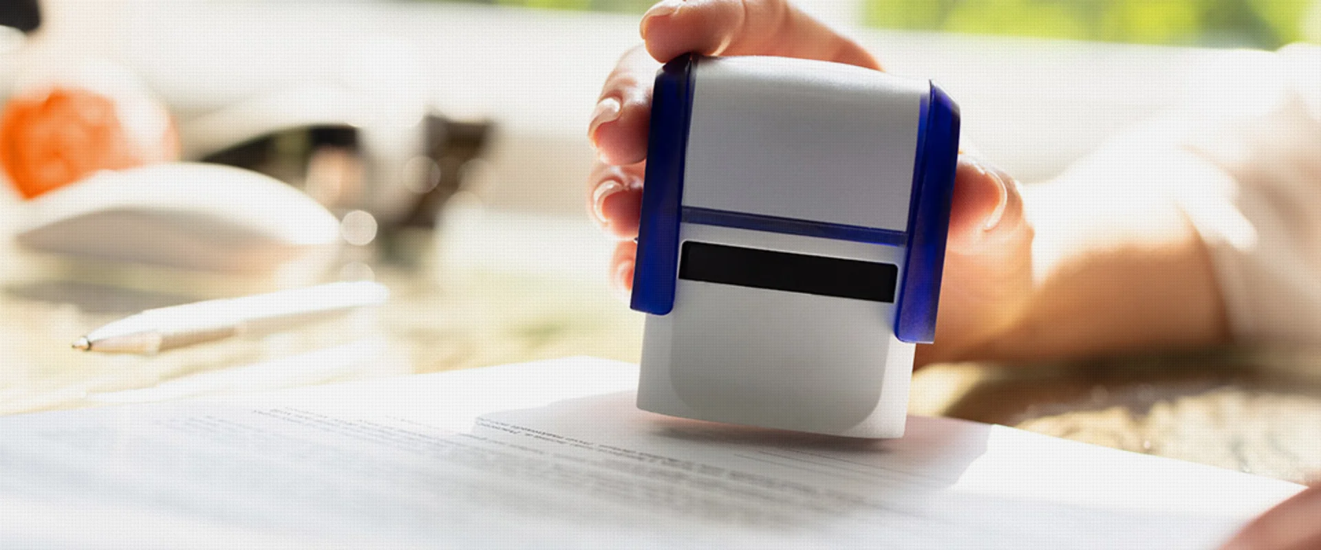 Eine Hand, die einen Stempel über einem Dokument hält, bereit, es zu stempeln, mit einem Stift und unscharfen Gegenständen im Hintergrund.