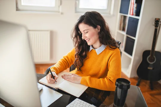 Eine Frau in gelbem Pullover schreibt in ein Notizbuch vor einem Computer, im Hintergrund eine Gitarre.