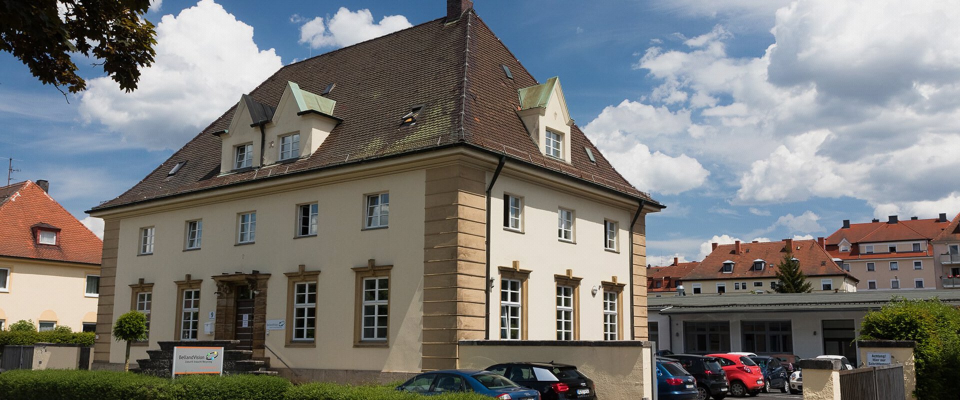 Ein historisches, zweistöckiges, sandfarbenes Gebäude mit einem dunklen Dach und heller Umrandung der Fenster zeigt den Unternehmensstandort von BellandVision in Pegnitz. 