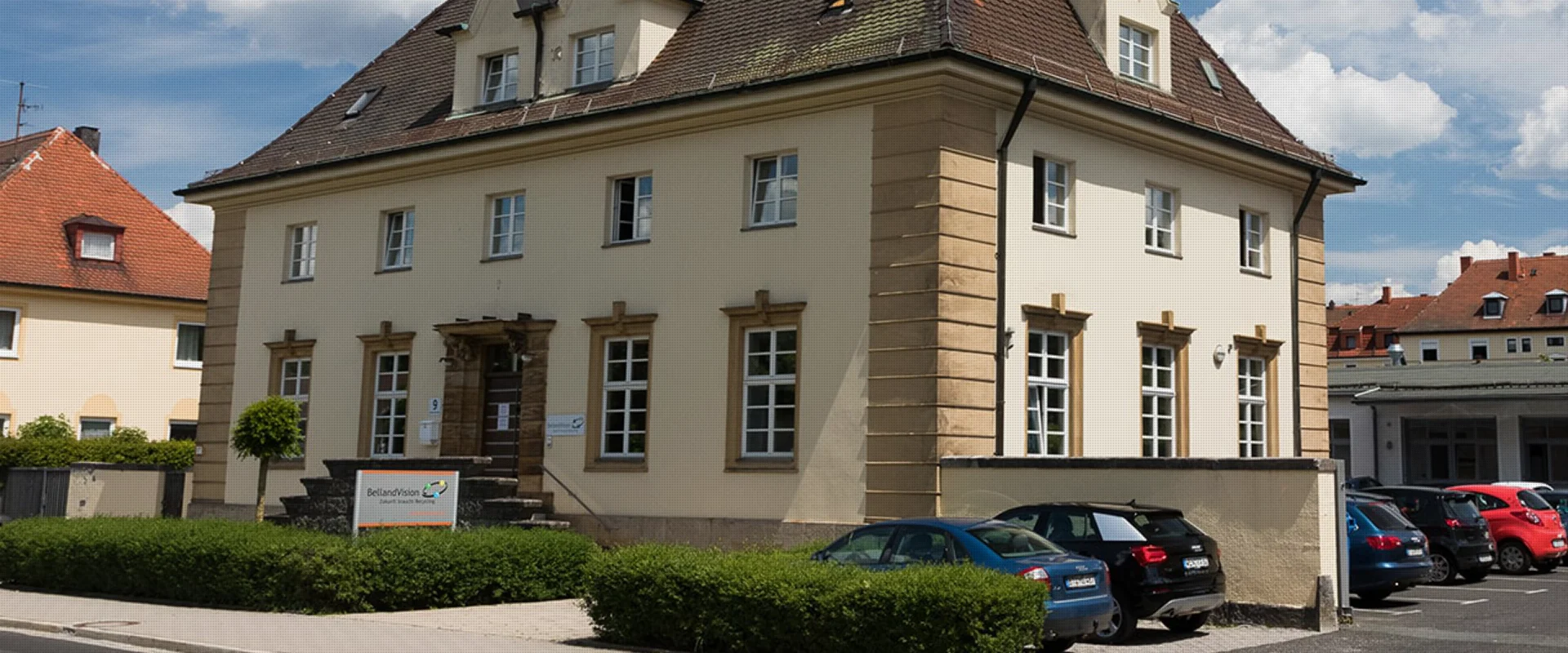 Ein historisches, zweistöckiges, sandfarbenes Gebäude mit einem dunklen Dach und heller Umrandung der Fenster zeigt den Unternehmensstandort von BellandVision in Pegnitz. 