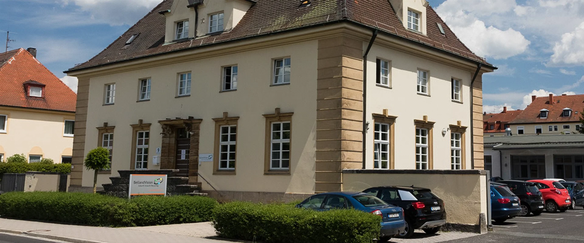 Ein historisches, zweistöckiges, sandfarbenes Gebäude mit einem dunklen Dach und heller Umrandung der Fenster zeigt den Unternehmensstandort von BellandVision in Pegnitz. 