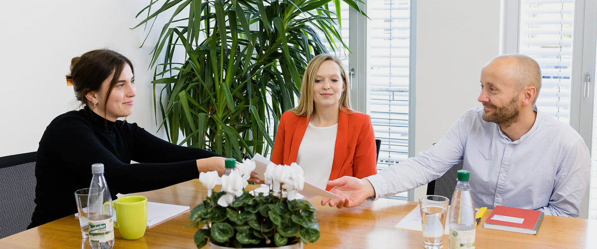 Drei Kollegen sitzen an einem Konferenztisch bei einem Meeting. Eine Frau links im Bild reicht Dokumente über den Tisch an einen Mann, der rechts sitzt.