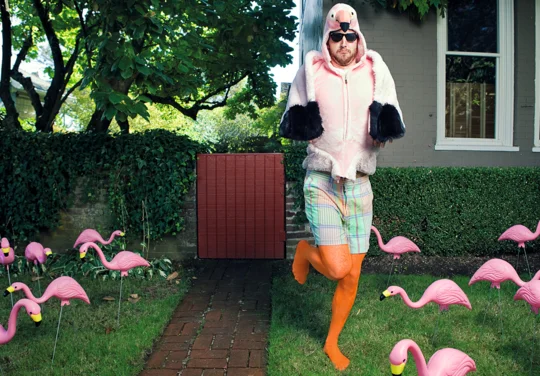 Mann in einem auffälligen Flamingo-Kostüm und Sonnenbrille steht in orangefarbenen Strümpfen und mit gehobenem Bein neben Plastikflamingos im Garten.