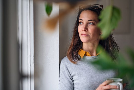 Eine Frau mit einer Tasse in der Hand schaut nachdenklich aus dem Fenster. Im Vordergrund sieht man verschwommen  Zimmerpflanzen.