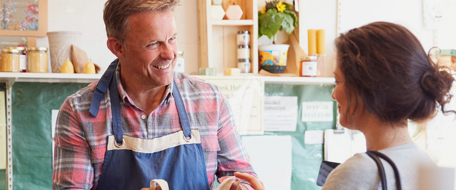 Ein lächelnder Verkäufer mit einer Schürze bedient eine Kundin in einem kleinen Lebensmittelladen.