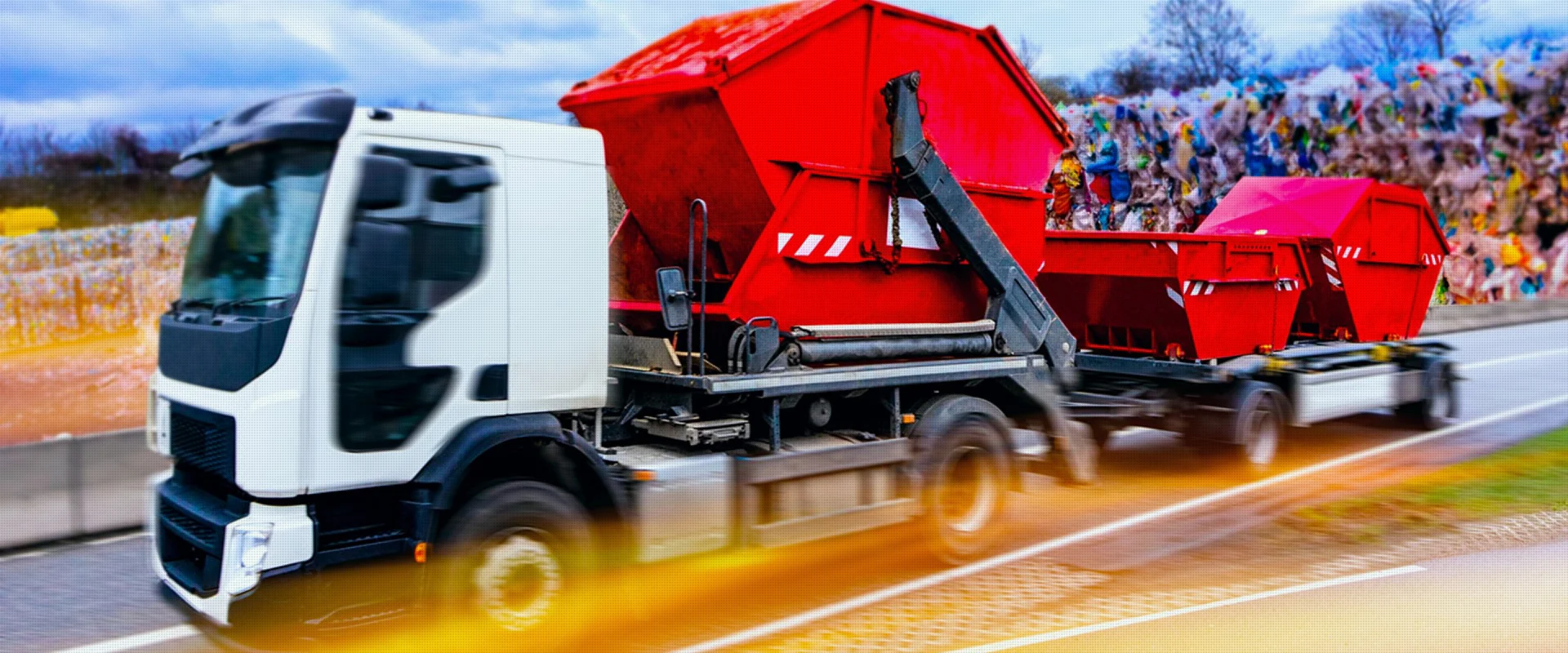 Ein  Müllentsorgungslastwagen mit roten Containern auf der Straße mit unscharfem Hintergrund, der eine Mülldeponie andeutet.