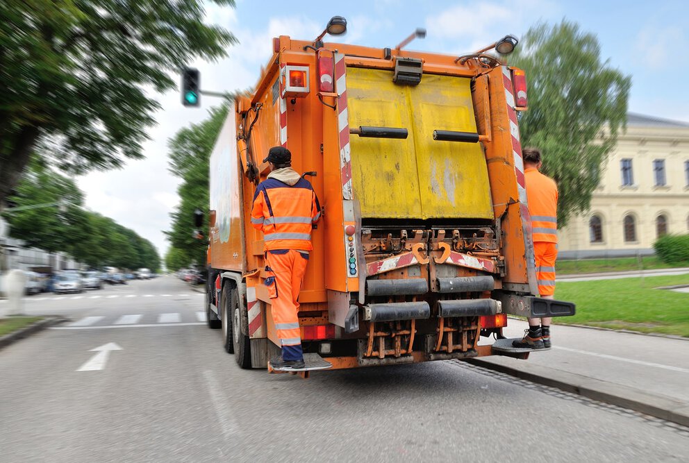 Zwei Müllwerker in Warnwesten auf  der Rückseite eines  orangefarbenen Müllwagens in einer Stadt.