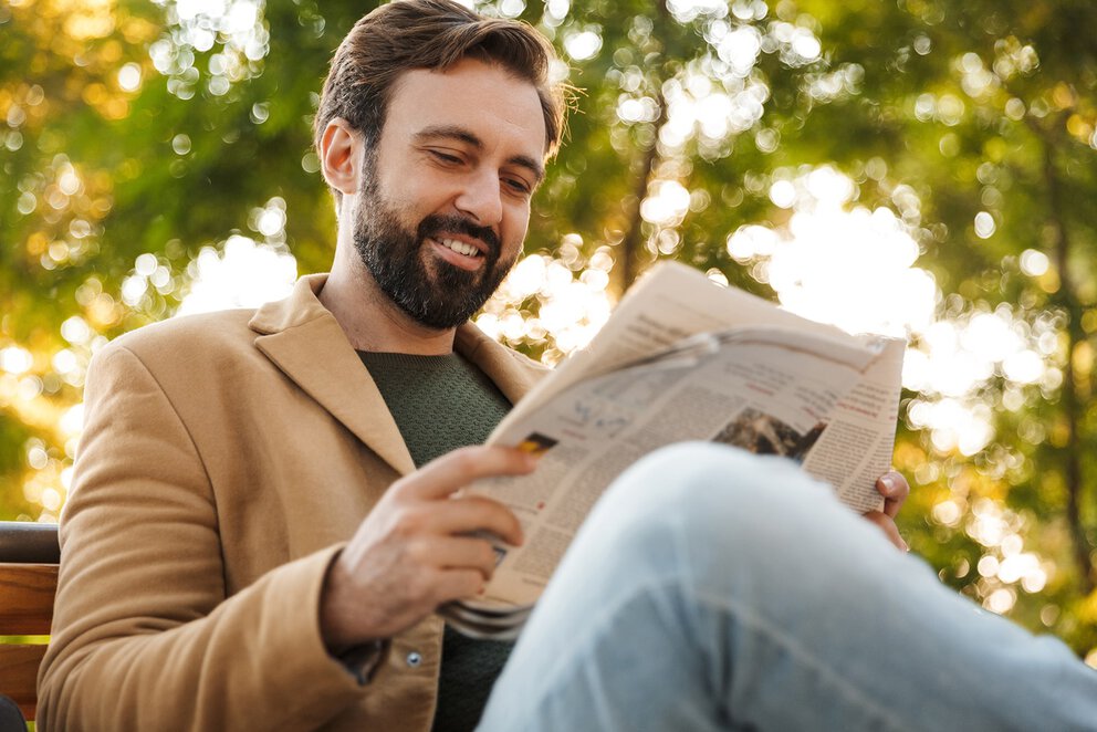 Lächelnder Mann mit dunklem Bart liest Zeitung im Freien mit sonnendurchfluteten Bäumen im Hintergrund.